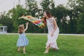 Smiling woman in light dress and little cute child baby girl play with colorful kite on grass in green park. Mother Royalty Free Stock Photo