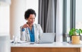 Smiling young adult mature African American businesswoman working on a laptop at her desk in a bright modern office holding her Royalty Free Stock Photo