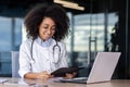 Smiling young African American female doctor working in clinic office with laptop and documents, sitting at table Royalty Free Stock Photo