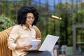 Smiling young African American female student sitting on a bench on a university campus studying online using a laptop Royalty Free Stock Photo