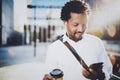Smiling young African American man in headphone walking at sunny city with take away coffee and using his mobile phone Royalty Free Stock Photo