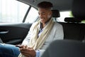 Smiling young african american man looking at mobile phone while in backseat of car