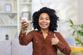 Smiling young African American woman sitting on sofa at home, holding air conditioner remote control pointing at wall Royalty Free Stock Photo