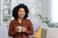 Smiling young African American woman sitting on sofa at home, holding cup in hands and looking away contentedly. Close Royalty Free Stock Photo