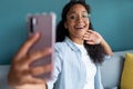 Smiling young african american woman using her mobile phone to take a selfie while sitting on sofa at home Royalty Free Stock Photo