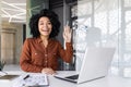 Smiling young African American woman working in the office using a laptop, sitting at the desk, waving and greeting at Royalty Free Stock Photo