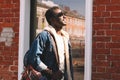 Smiling young african man wearing a jeans jacket and backpack looking up at sunlight while walking on a city street over Royalty Free Stock Photo