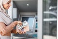 Smiling young arab islamic business woman in hijab puts dollars in wallet near atm with blank screen Royalty Free Stock Photo