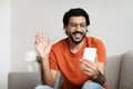 Smiling young arab male in glasses sits on couch, waving hand, has video call on smartphone in living room