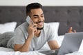 Smiling young arab man talking on cellphone and using laptop in bed Royalty Free Stock Photo