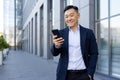 Smiling young asian business man lawyer standing outside office center and using mobile phone, hand in pocket Royalty Free Stock Photo