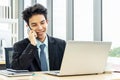 Smiling young asian businessman sitting at desk  talking on mobile phone and using laptop in workplace Royalty Free Stock Photo