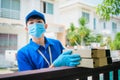 A smiling young asian delivery in blue uniform with parcel cardboard in front of customer house