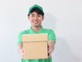 Smiling young Asian delivery man in green T-shirt uniform and cap holding cardboard box on white background, giving box on camera