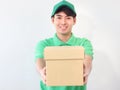 Smiling young Asian delivery man in green T-shirt uniform and cap holding cardboard box on white background, giving box on camera