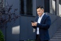 A smiling young Asian male businessman is standing on a city street near a business building, holding and using a mobile Royalty Free Stock Photo