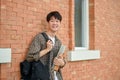 A smiling young Asian male college student stands by a brick wall on campus, carrying a backpack