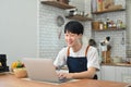 Smiling young asian man freelancer using laptop, working remote job from home while sitting in cozy kitchen interior Royalty Free Stock Photo