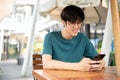 A smiling young Asian man wearing glasses sits at a wooden table outdoors, looking at his smartphone Royalty Free Stock Photo