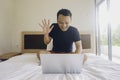 Smiling young Asian man works remotely behind a laptop at home, sitting in bed Royalty Free Stock Photo
