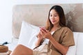 Smiling young Asian traveler woman sitting and using mobile phone on bed in hotel room. Travel alone, summer and holiday concept Royalty Free Stock Photo