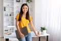 Smiling young asian woman holding closed laptop, standing in well-lit home office Royalty Free Stock Photo