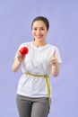 Smiling young asian woman holding measuring tape around her waist and eating red apple over violet background Royalty Free Stock Photo