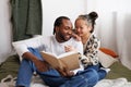 Smiling young asian woman hugging african american boyfriend with book on bed at home Royalty Free Stock Photo