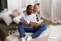 Smiling young asian woman hugging african american boyfriend reading book near laptop on bed Royalty Free Stock Photo
