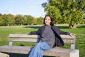 Smiling young asian woman in outdoor jacket, sitting on bench in green sunny park, resting alone, relaxing on fresh air Royalty Free Stock Photo