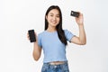 Smiling young asian woman showing credit card in raised hand, empty phone screen, concept of online banking and shopping Royalty Free Stock Photo