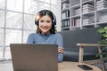 Smiling young asian woman using laptop web camera while wearing headphones and sitting on the rug beside to the sofa at Royalty Free Stock Photo