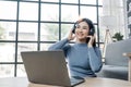 Smiling young asian woman using laptop web camera while wearing headphones and sitting on the rug beside to the sofa at Royalty Free Stock Photo