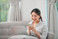 Smiling young asian woman using mobile phone while sitting on a couch at home with laptop computer Royalty Free Stock Photo