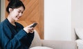 Smiling young asian woman using mobile phone while sitting on a couch at home with laptop computer Royalty Free Stock Photo