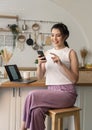 Smiling young asian woman using mobile phone while sitting in kitchen room at home with laptop computer Royalty Free Stock Photo