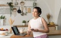 Smiling young asian woman using mobile phone while sitting in kitchen room at home with laptop computer Royalty Free Stock Photo