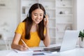 Smiling, young asian woman wearing headphones taking notes and using her laptop Royalty Free Stock Photo