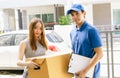 Smiling young Asian woman receiving package box from Asian delivery man in blue uniform at front of house and looking at camera.