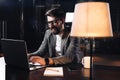 Smiling young bearded businessman working on contemporary notebook in loft office at night Royalty Free Stock Photo