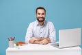 Smiling young bearded man in light shirt sit, work at white desk with pc laptop isolated on pastel blue background Royalty Free Stock Photo