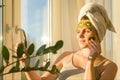 Smiling young beautiful woman close up at home near the window with natural homemade fruit facial mask of kiwi on face, towel on Royalty Free Stock Photo