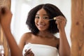 Smiling young black lady in towel using facial serum near mirror, taking care about her beauty at home Royalty Free Stock Photo