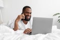 Smiling young black man with beard speaks by phone, looks at laptop on white bed in bedroom interior Royalty Free Stock Photo