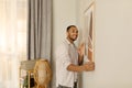 Smiling young black man putting picture frame, hanging painting on wall. Home interior and domestic decor Royalty Free Stock Photo