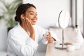 Smiling young black woman in white bathrobe applying skincare product Royalty Free Stock Photo
