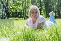 A smiling young blonde woman lies on the grass in a park with a laptop on a summer sunny day. Blogging, online communication,