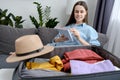 Smiling young brunette girl sitting on comfy couch in living room at home writing travel checklist while packing clothes to Royalty Free Stock Photo