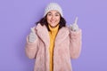 Smiling young brunette woman dresses in yellow sweater, pink faux fur coat and cap, posing isolated on lilac background, pointing