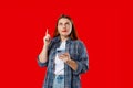 Smiling young brunette woman with smart phone pointing with the index finger a great idea on red background, studio Royalty Free Stock Photo